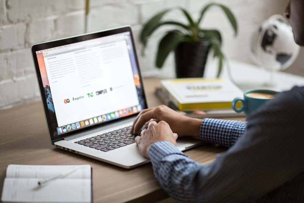 A person sitting at a desk using a laptop, with a cup of coffee and a notebook nearby. The laptop screen displays a webpage with various logos and text with website copywriting for professional services